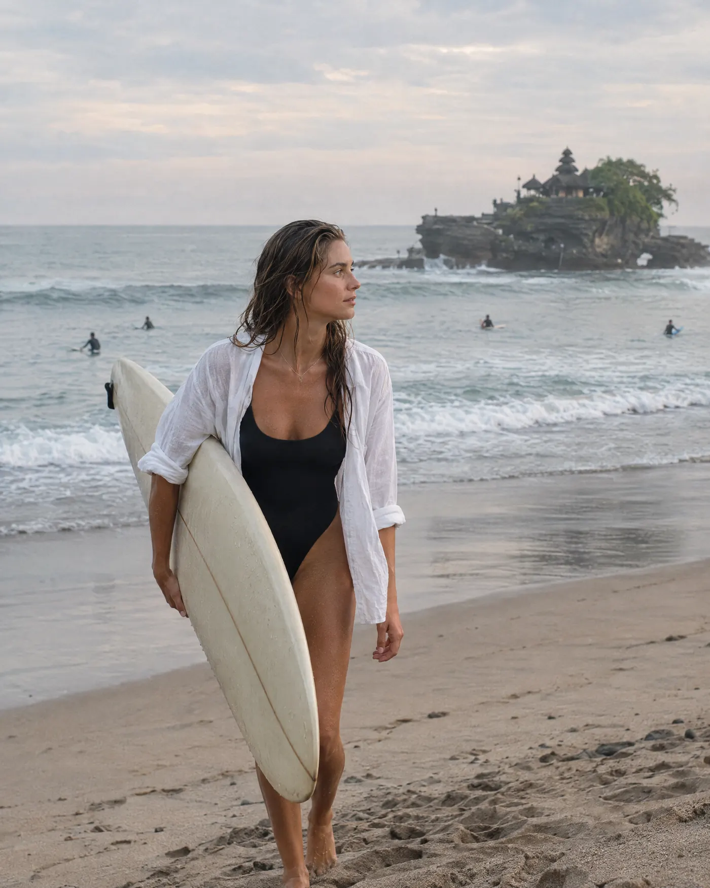 A surfer walking up the beach with her board near a sea temple in Bali, early morning